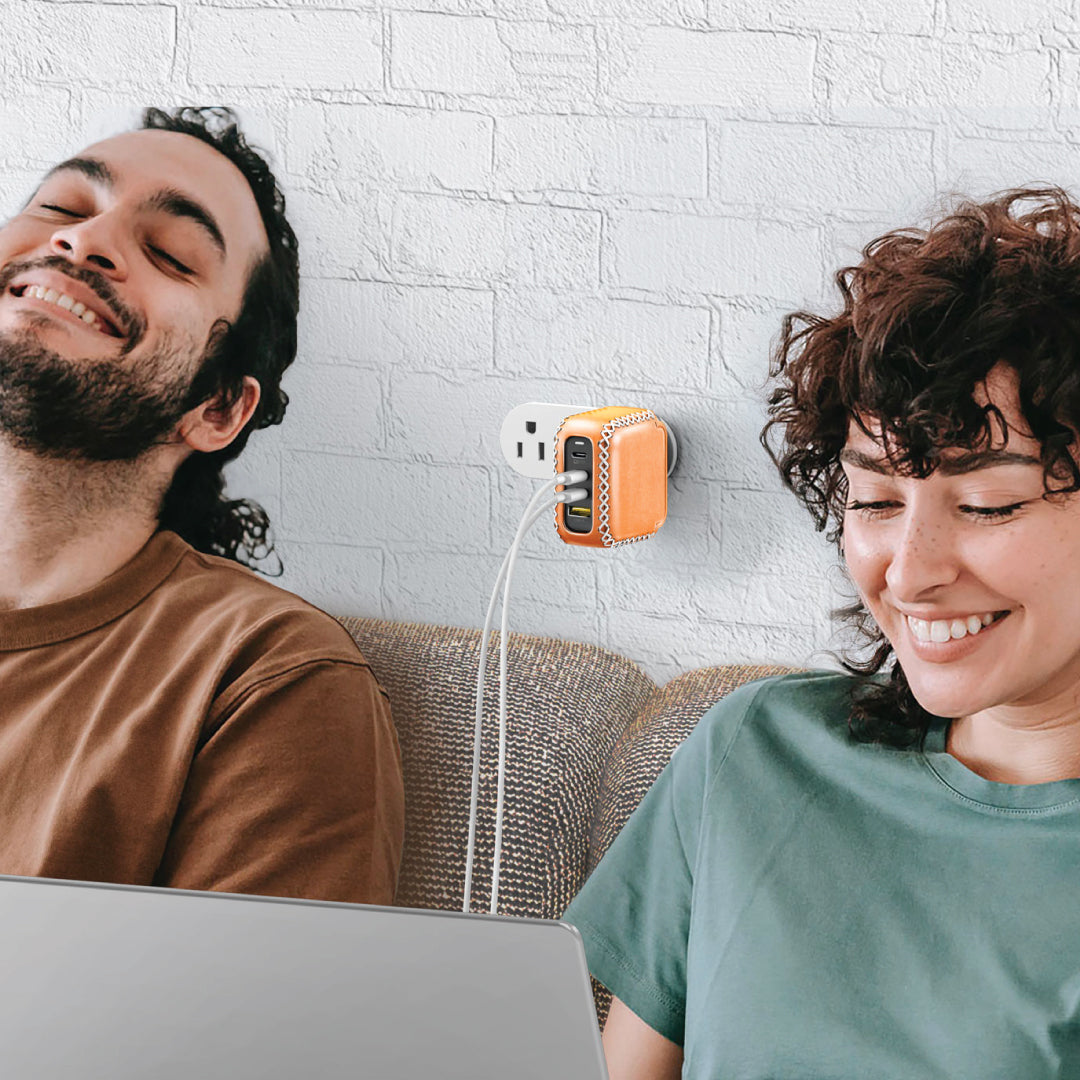 Two people sitting on a couch with a laptop, charing laptops with a VogDUO 100W leather-design wall charger smiling and looking at something off-camera.