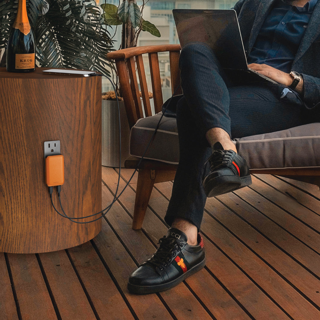 Person sitting wearing suits sittingon a chair with a laptop, wearing black sneakers, using VogDUO leather design wall charger on his digital nomad trip a wooden deck.