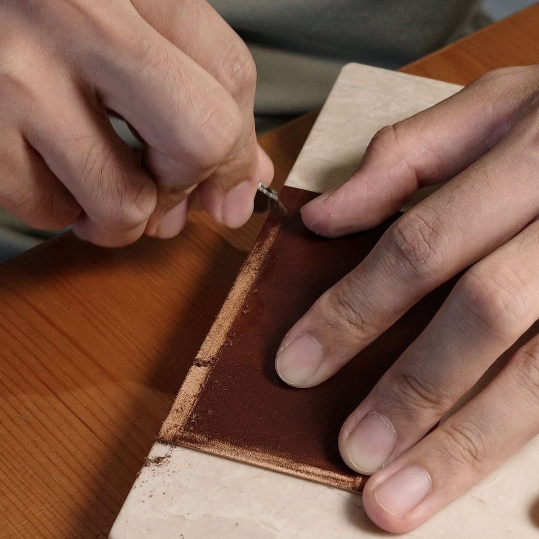 Close-up of hands working on a leather item with a peeling tool on a wooden surface.