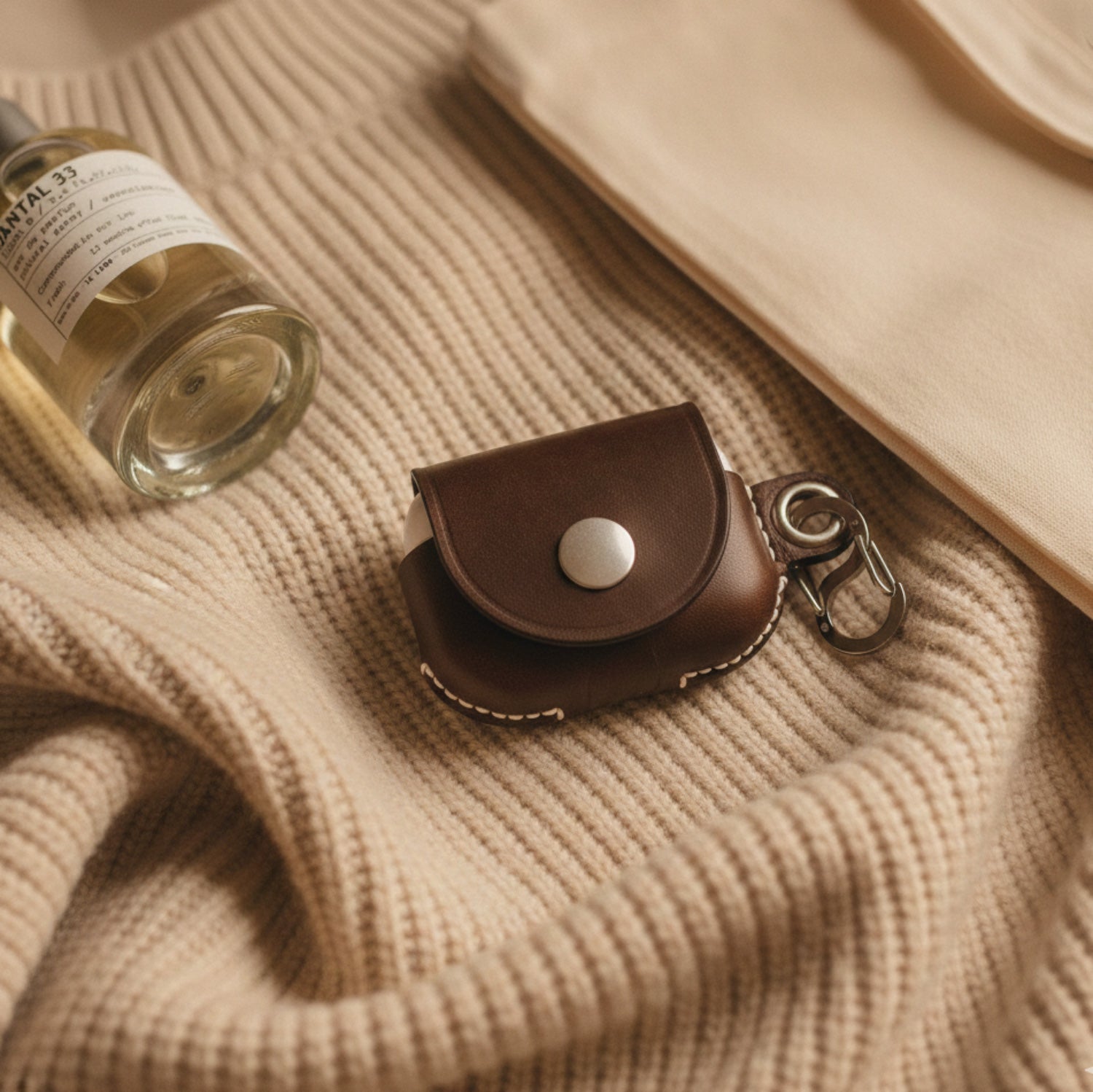 A top-down, first-person perspective shot featuring a VogDUO dark brown leather AirPods Pro 3 case resting on a thick, ribbed oatmeal-colored knit sweater. To the left, a glass bottle of Le Labo Santal 33 perfume is partially visible, and to the right, a cream-colored cotton tote bag adds a soft layer to the composition. The lighting is warm and soft, emphasizing the rich grain of the leather against the heavy texture of the knit fabric.