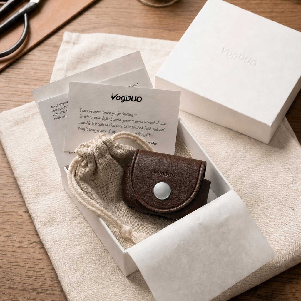 A small brown leather card holder for AirPods Pro 3 with a silver snap button sits in a white gift box with a beige drawstring pouch, branded VogDUO, on a wooden table.