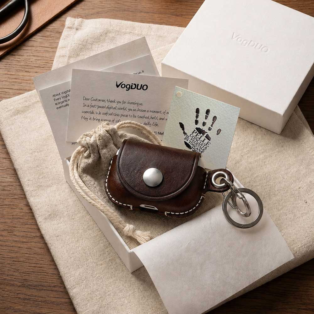 A small brown leather AirPods Pro case with a silver snap button and metal key ring, presented in a white box with branded cards and a dust bag. The box lid and packaging are branded VogDUO.