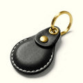 A black leather keychain case for AirTag with white contrast stitching, a gold snap button, and a gold split key ring, shown on a clean off-white background.
