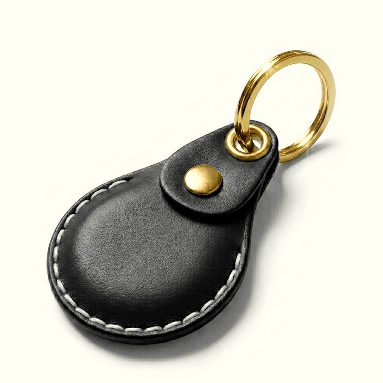 A black leather keychain case for AirTag with white contrast stitching, a gold snap button, and a gold split key ring, shown on a clean off-white background.