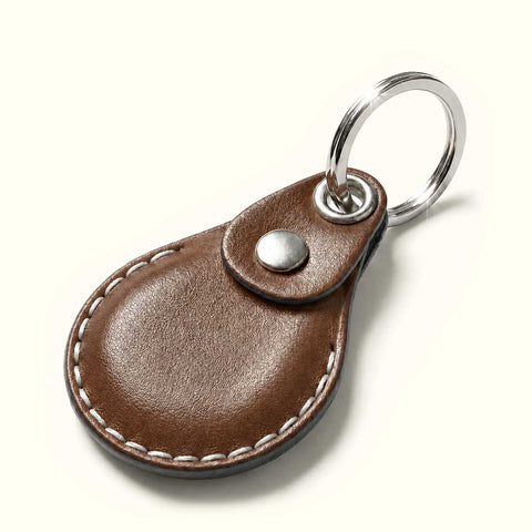 A brown leather keychain design for AirTag with a teardrop-shaped body, white contrast stitching, a snap button closure, and a silver metal key ring on a light cream background.