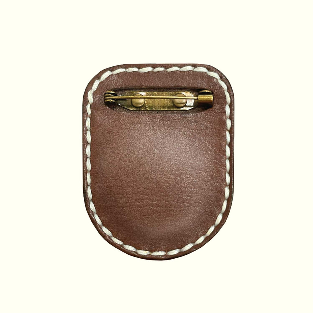 A brown leather brooch-pin holder for AirTag with white contrast stitching and a brass pin clasp