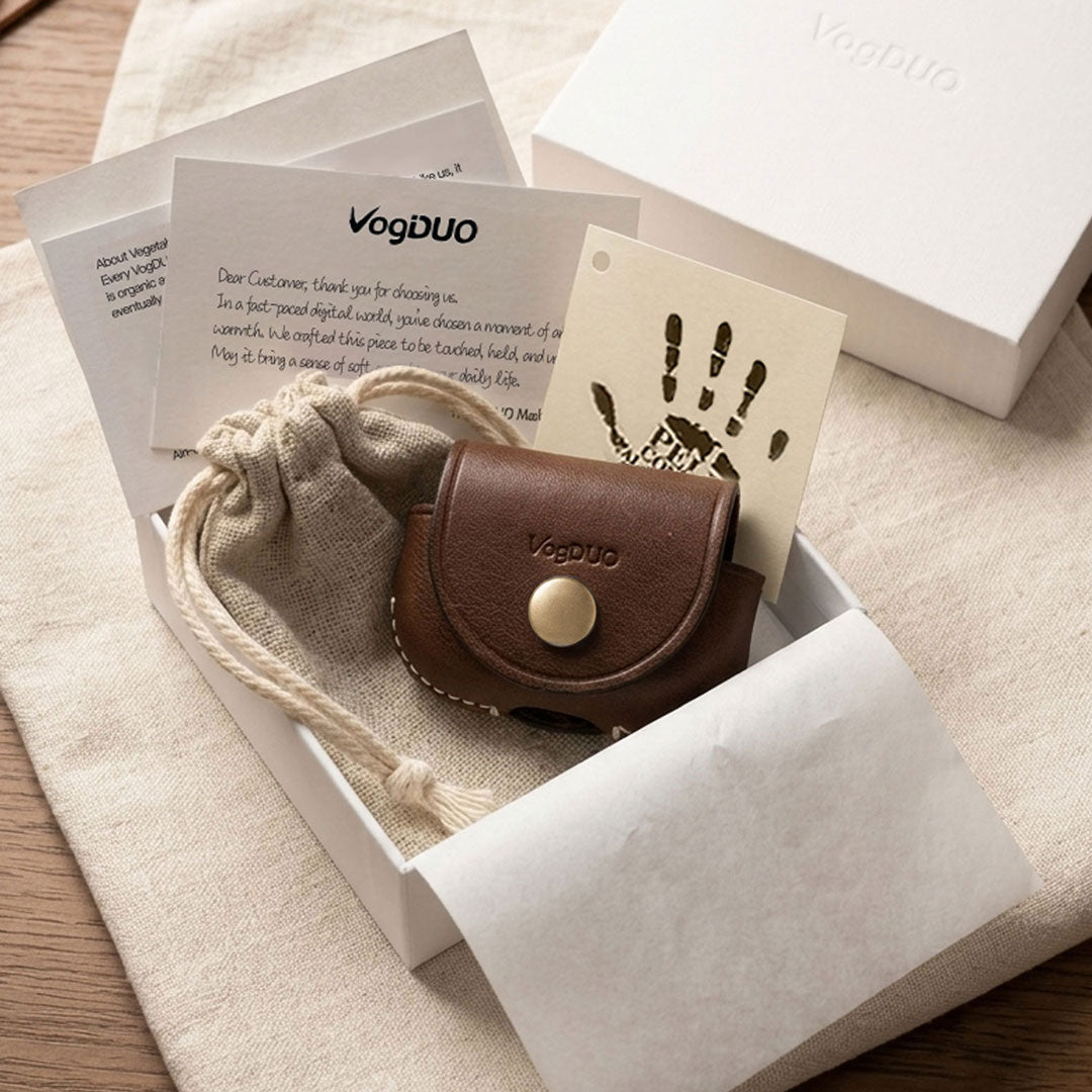 A small brown leather AirPods Pro Case with a snap closure and embossed VegDUO logo, presented in a white box with a beige drawstring pouch, cards, and a branded box on a neutral fabric surface.
