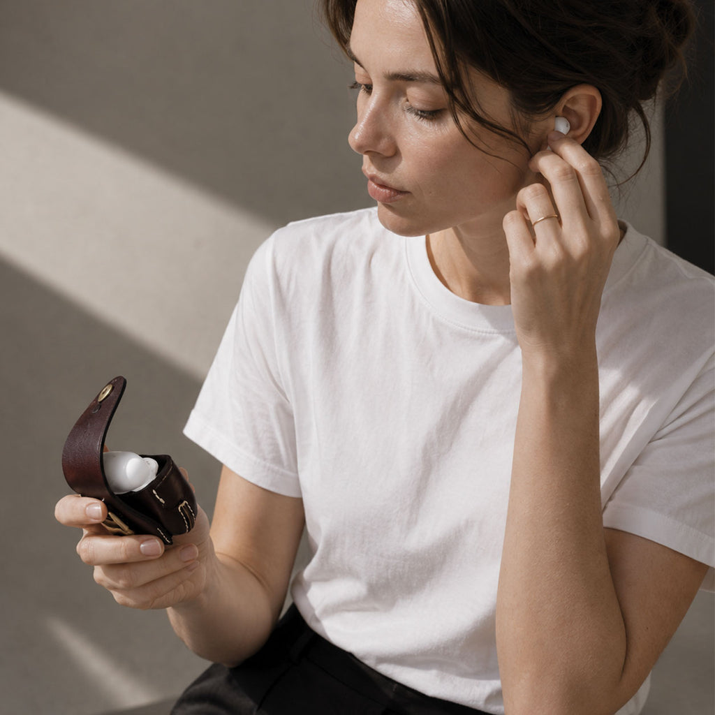 A woman in a white t-shirt is shown in a lifestyle setting, holding an open dark brown leather AirPods Pro 3 case and inserting a white wireless earbud into her ear. The background is a clean, neutral gray wall with soft diagonal shadows.