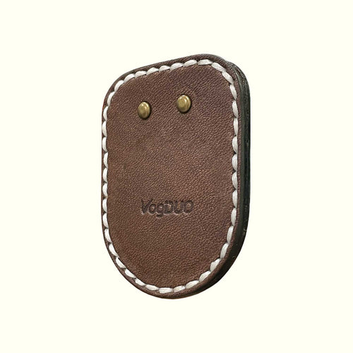 A brown leather case for AirTag with white stitching and brass rivets, featuring the embossed brand name 'VogDUO' on a light cream background.