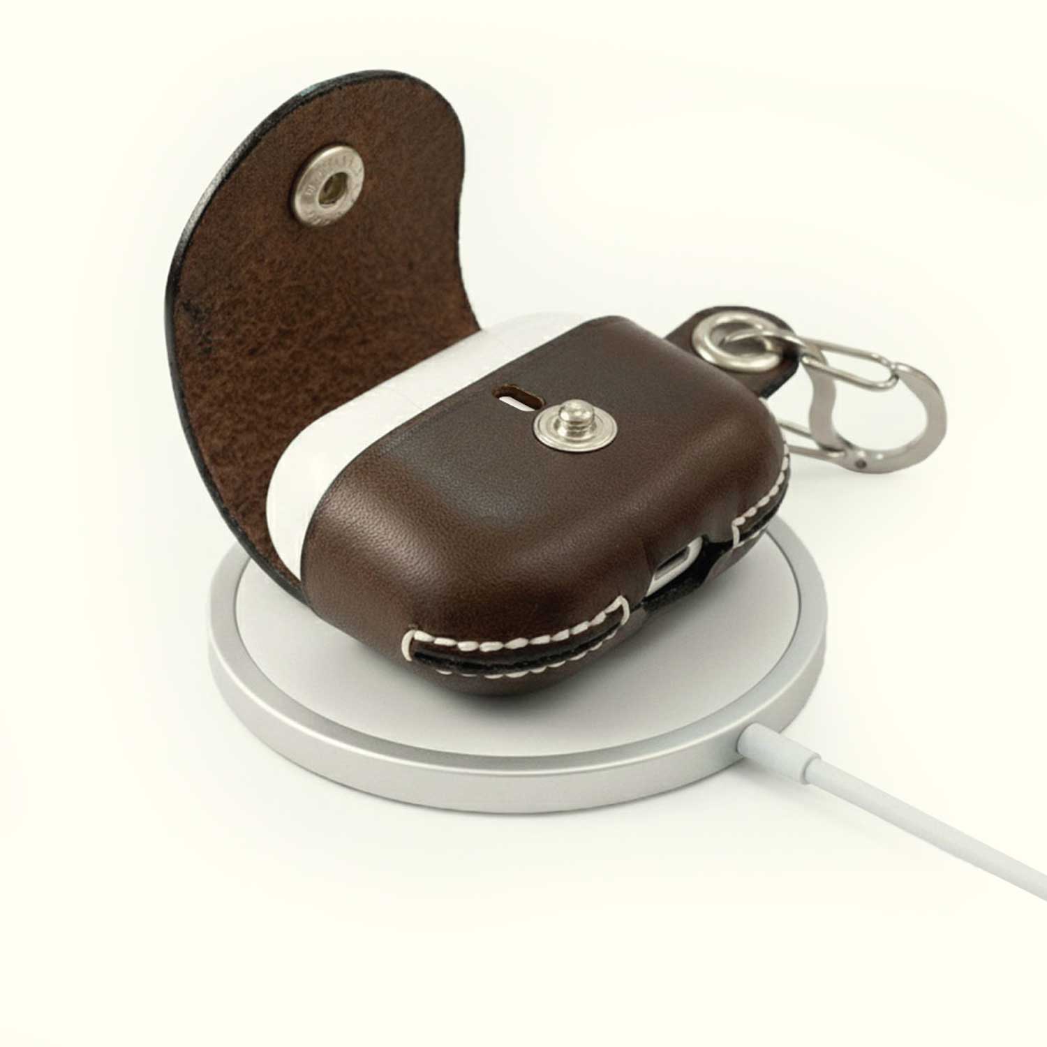 A brown leather AirPods case cover with a snap-button closure and keychain clip, shown open with a white AirPods case inside, resting on a silver wireless charging pad against a white background.