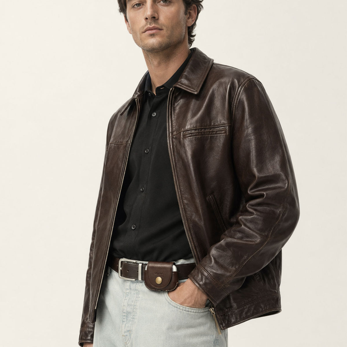 A man wearing a dark brown leather jacket over a black button-down shirt and light blue jeans with an AirPods Pro 3 Belt-clip case, photographed against a plain off-white studio background.