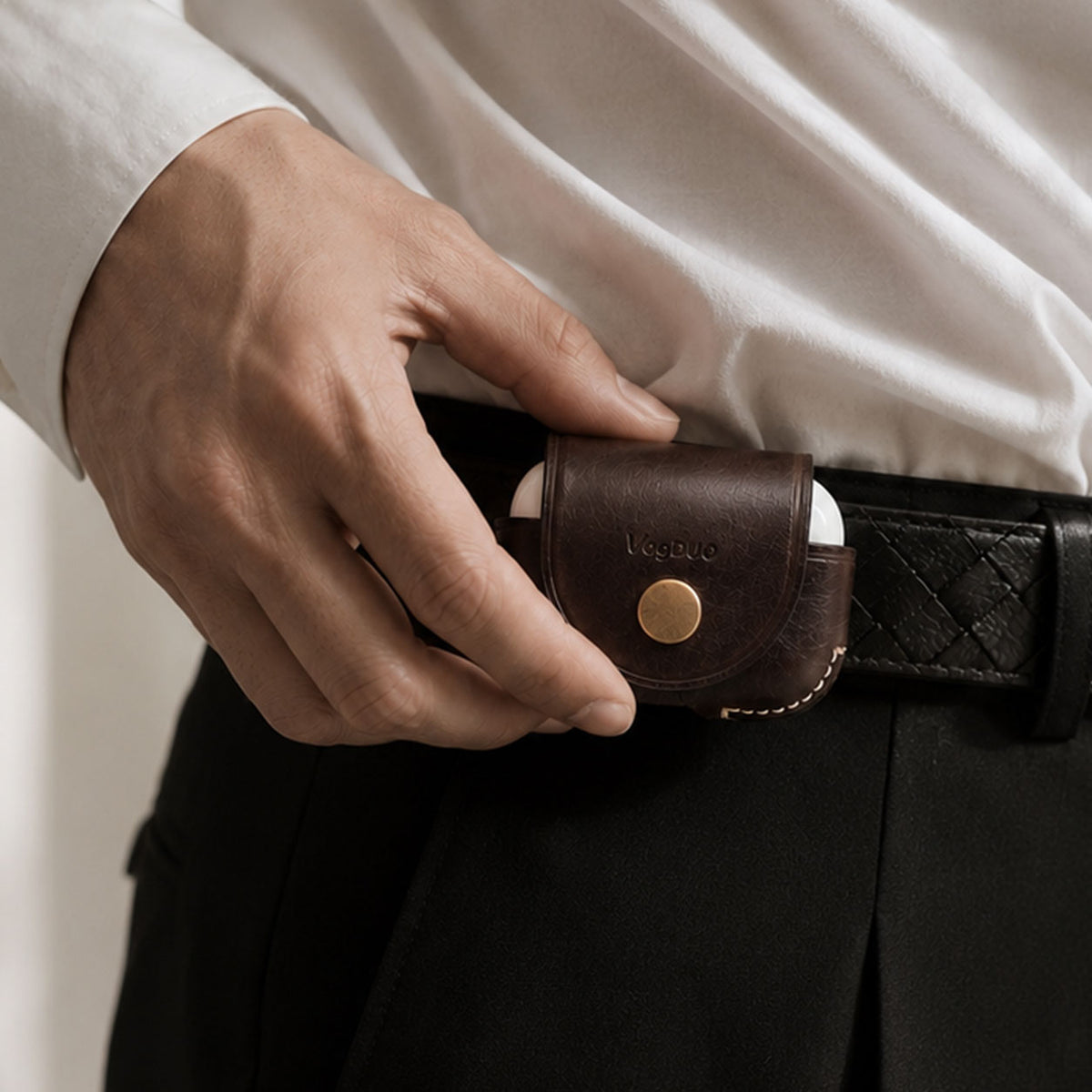 A close-up of a person's hand inserting a brown leather AirPods Pro case into a black belt pouch at the waist, wearing a white shirt and black trousers.