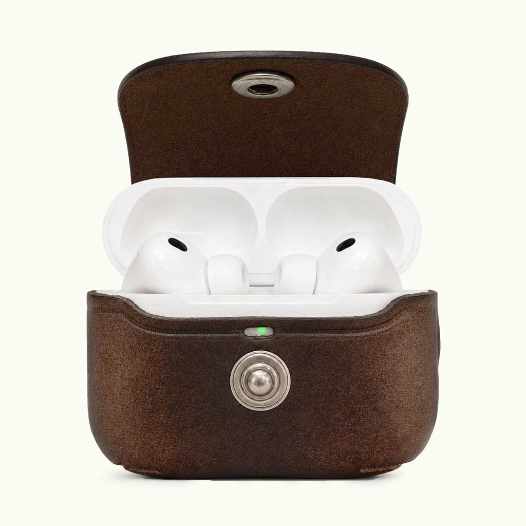 A brown textured leather AirPods Pro 3 charging case shown open, revealing white earbuds inside. The case has a silver metal button on the front and a small green LED indicator light, set against a plain off-white background.