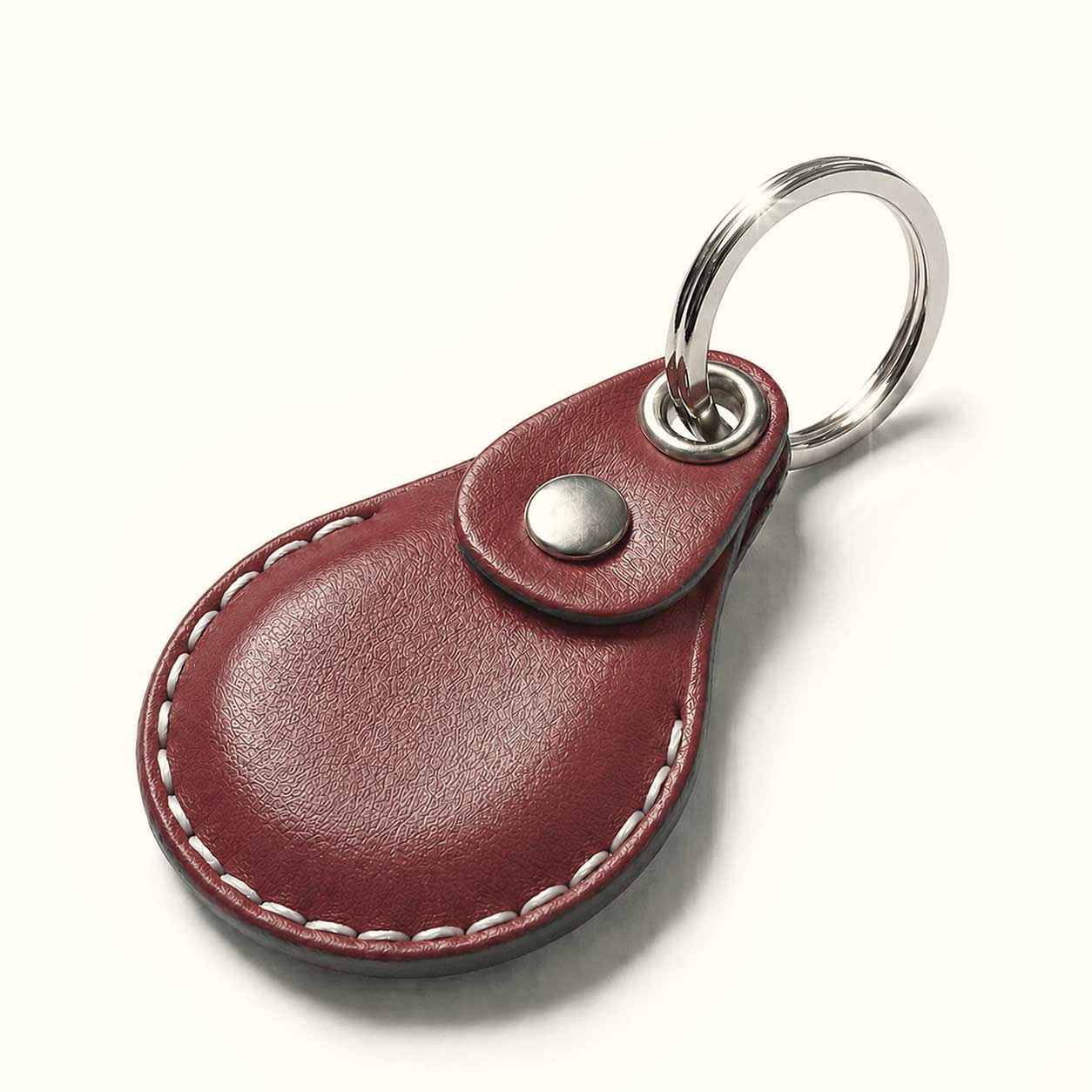 A teardrop-shaped wine-red leather keychain for AirTag with white contrast stitching, a snap button closure, and a silver metal key ring, shown on a light cream background.