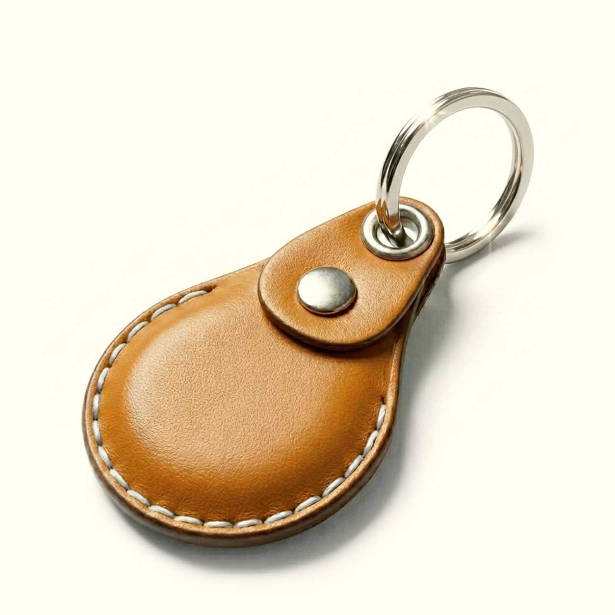 A tan leather keychain case for AirTag with a teardrop-shaped body, white contrast stitching, a snap button closure, and a silver metal key ring, shown on a clean white background.