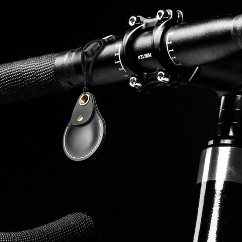 Close-up of a black AirTag leather keychain attached to a bicycle handlebar with a black background.