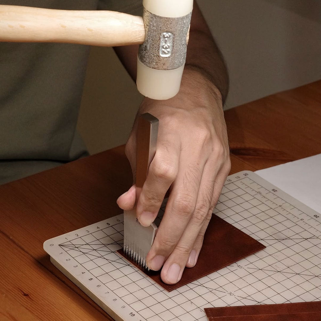 Hand using a French Chisel on a piece of leather to locate the stitches on a cutting mat with grid lines.