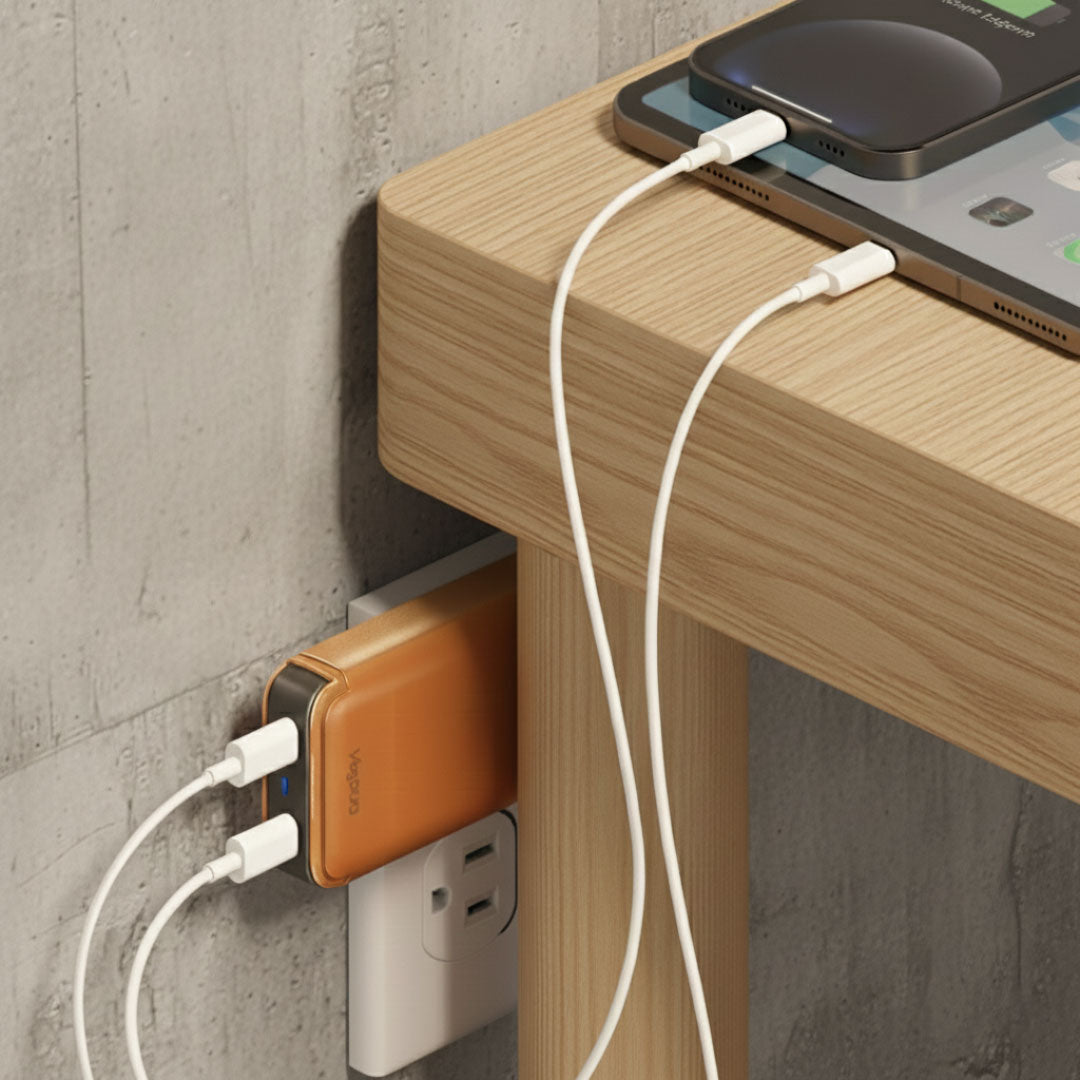 Smartphone charging on a wooden surface with a VogDUO ultra-slim wall charger plugged into an outlet behind a table, and in the gap.