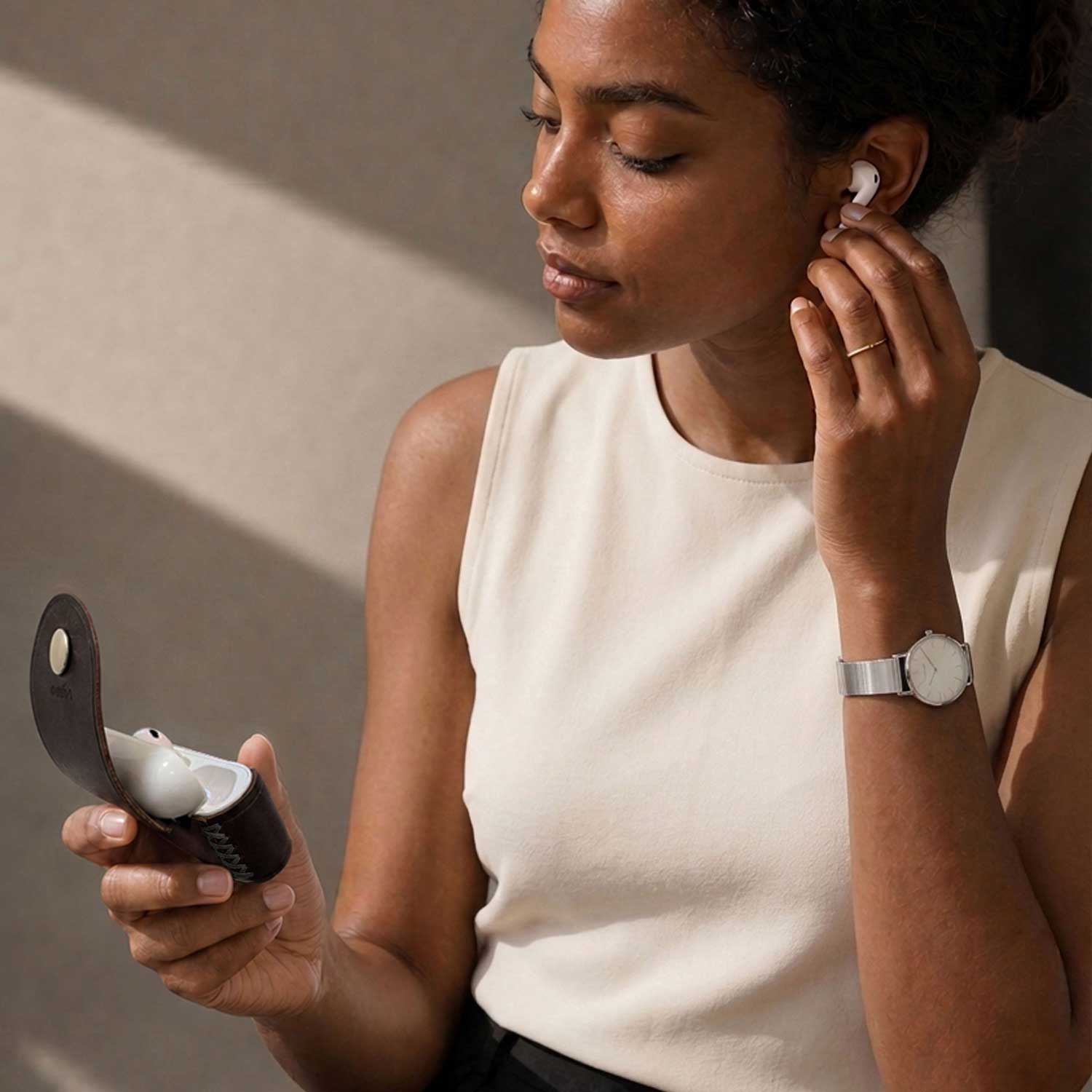 A woman wearing a AirPods Pro 3 earbud and holding its charging case, with a matching white earbud resting in the genuine leather case.