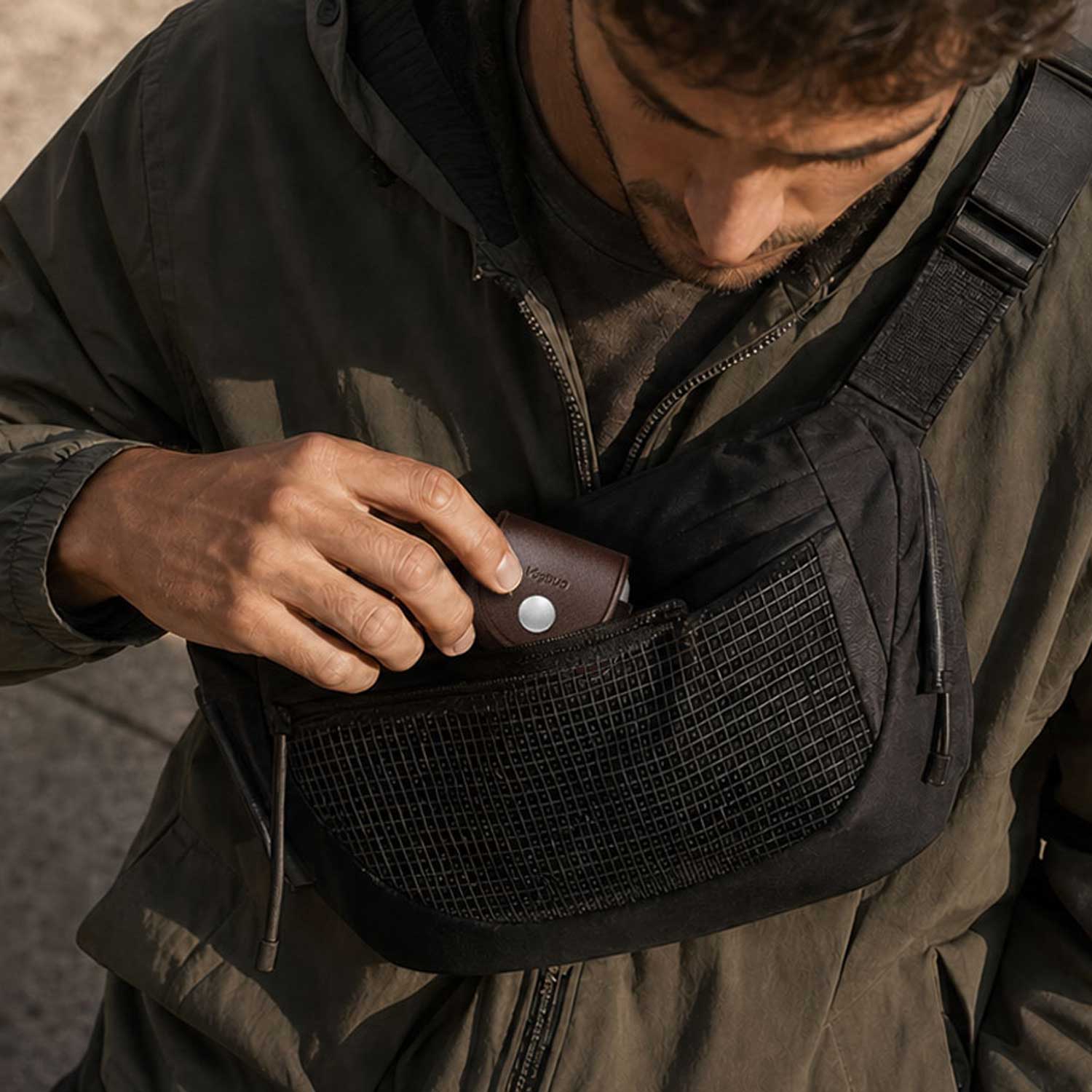 A close-up of a man wearing a black crossbody sling bag with a mesh pocket, shown as he inserts a brown leather AirPods Pro 3 case into the front pouch of the bag.