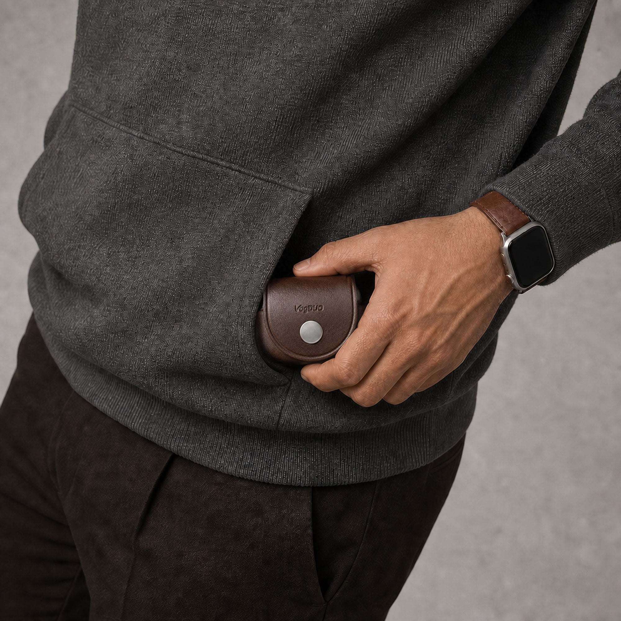 A close-up of a person wearing a smart casual dark gray knit sweater and brown pants, reaching into a side pocket to pull out a small brown leather case for AirPods Pro 3 with a silver button. The person is also wearing a smartwatch with a brown leather strap.